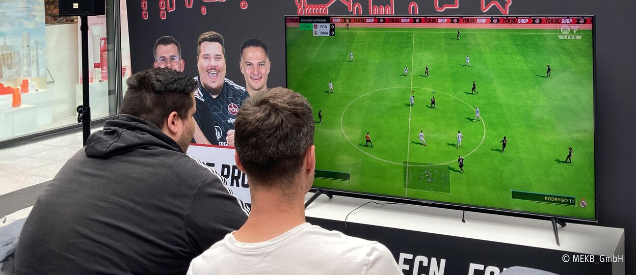Zwei junge Männer die im Hbf Nürnberg vor einem Fernseher sitzen und EA Sports FC gegeneinander spielen