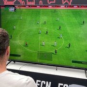 Zwei junge Männer die im Hbf Nürnberg vor einem Fernseher sitzen und EA Sports FC gegeneinander spielen