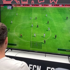 Zwei junge Männer die im Hbf Nürnberg vor einem Fernseher sitzen und EA Sports FC gegeneinander spielen