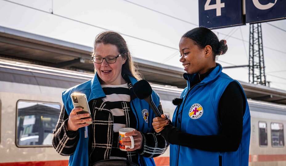 Zwei Frauen in blauen Bahnhofsmissions-Westen stehen am Bahnsteig und schauen ins Handy einer der Frauen.