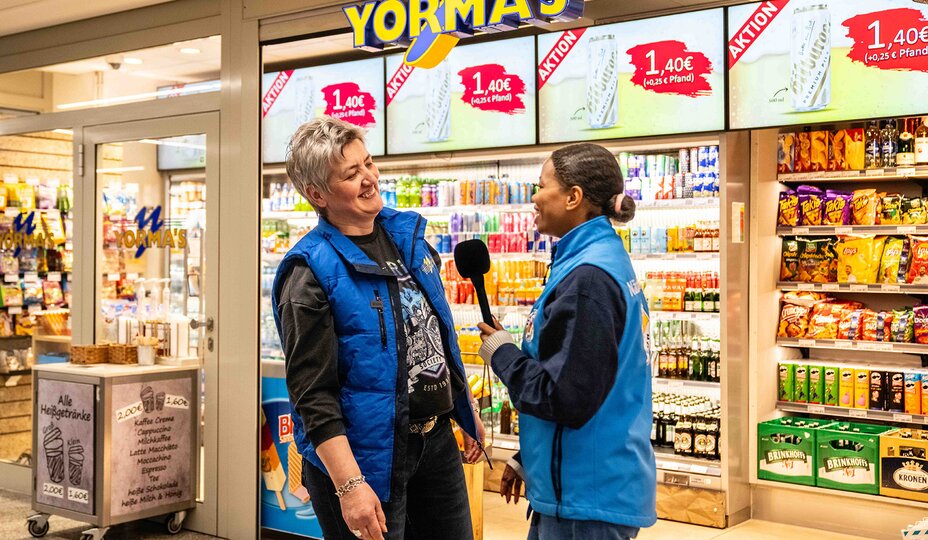 Eine junge Frau in blauer Bahnhofsmissions-Weste interviewt eine Frau vor einem Yorma's-Geschäft im Bahnhof.